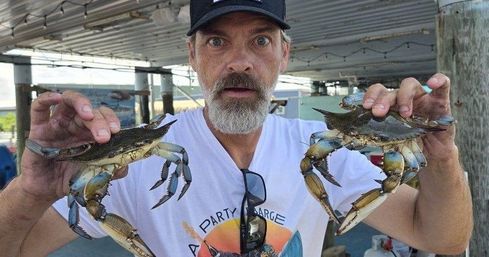 Wide-eyed man holding two live blue crabs by their shells at a covered marina dock, close-up showing crab claws and coastal seafood catch.