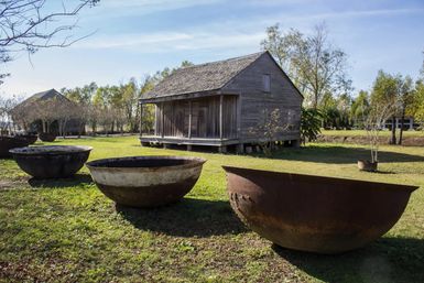 Giant rusty iron cauldrons on a grassy rural yard in front of a weathered wooden farmhouse beneath a clear blue sky