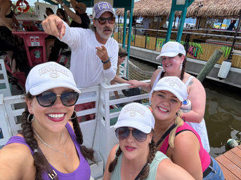 Group selfie of four smiling women in matching white caps and braided hair on a docked party boat, a man pointing behind them, tiki-style waterfront bars and water in the background.