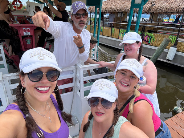 Group selfie of four smiling women in matching white caps and braided hair on a docked party boat, a man pointing behind them, tiki-style waterfront bars and water in the background.