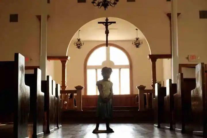 Sunlit church interior with wooden pews and a small child statue silhouette standing in the center aisle beneath a hanging crucifix and arched window.