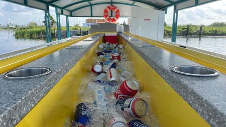 Yellow center cooler filled with ice and assorted soda and beer cans on a covered waterfront party boat, life preserver overhead and marshy shoreline and marina in the background