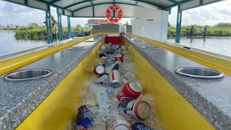 Yellow center cooler filled with ice and assorted soda and beer cans on a covered waterfront party boat, life preserver overhead and marshy shoreline and marina in the background