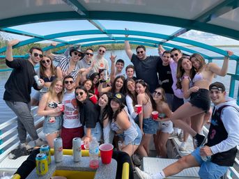 Smiling group of young adults on a turquoise‑canopied boat over a calm waterway, wearing swimsuits and summer clothes, holding red cups and cans — lively summertime boat party with marshy shoreline in the background.
