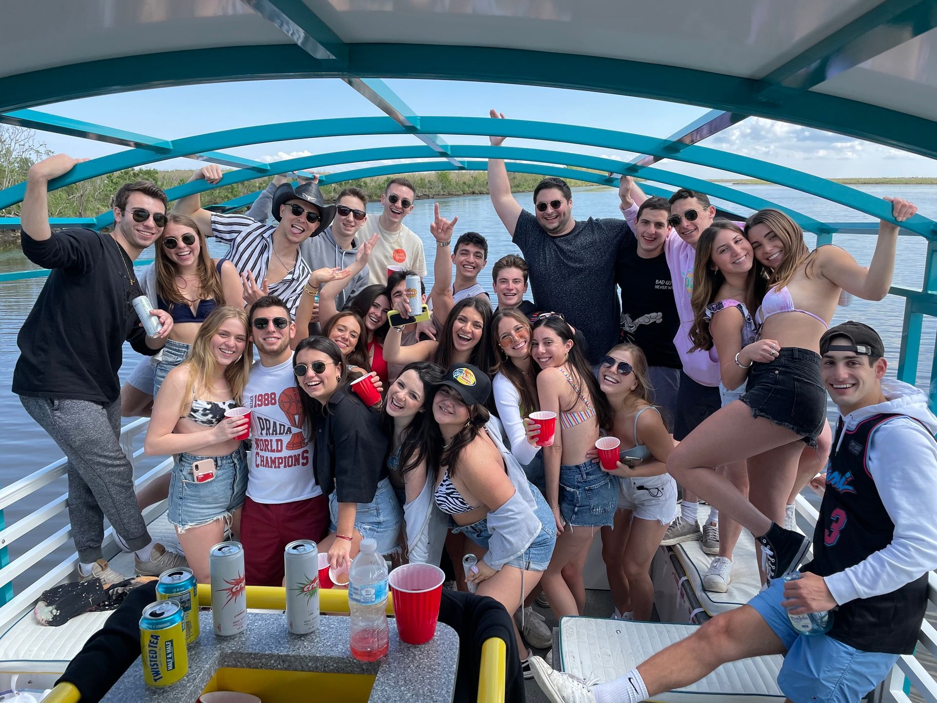 Group of young adults enjoying a sunny pontoon boat party on calm lake, wearing swimsuits and summer outfits, holding red cups and canned drinks under a turquoise canopy with marshland in the background.