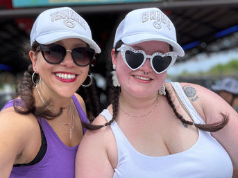 Two smiling women with braided pigtails wearing white caps at an outdoor bachelorette celebration — one in a purple tank with dark sunglasses, the other in a white tank wearing heart-shaped 'Bride to Be' sunglasses.