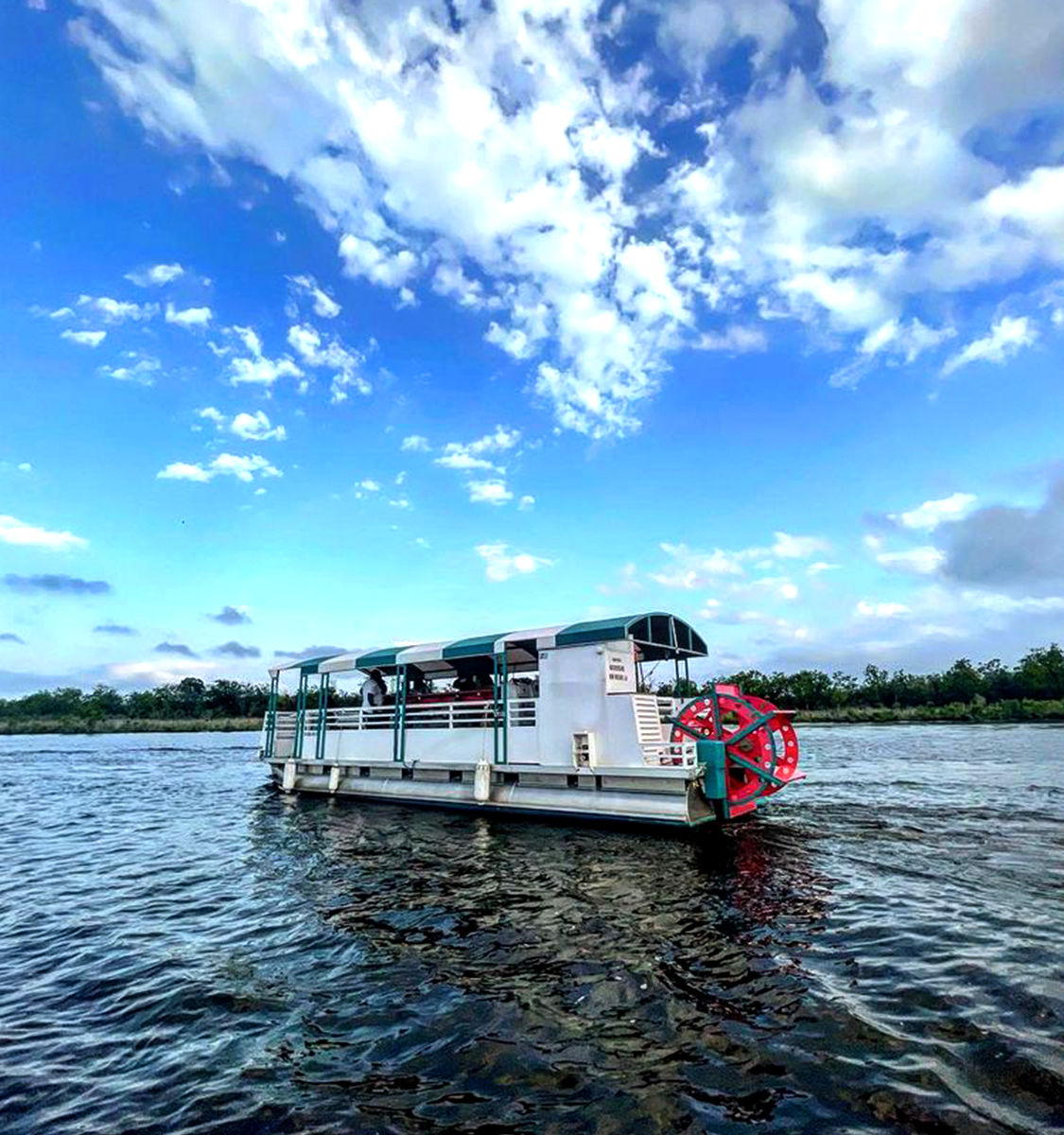 Pontoon-style paddlewheel riverboat with red wheel cruising a wide river under a bright blue sky and scattered clouds, tree-lined shore in the background