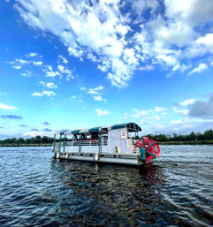 Pontoon-style paddlewheel riverboat with red wheel cruising a wide river under a bright blue sky and scattered clouds, tree-lined shore in the background