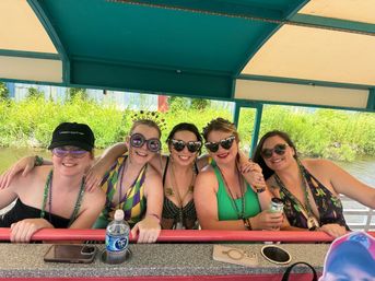 Five friends smiling on a covered riverboat cruise wearing colorful swimsuits, Mardi Gras beads and festive sunglasses, leaning on the railing with canal greenery behind them.