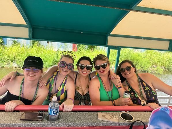 Five friends smiling on a covered riverboat cruise wearing colorful swimsuits, Mardi Gras beads and festive sunglasses, leaning on the railing with canal greenery behind them.