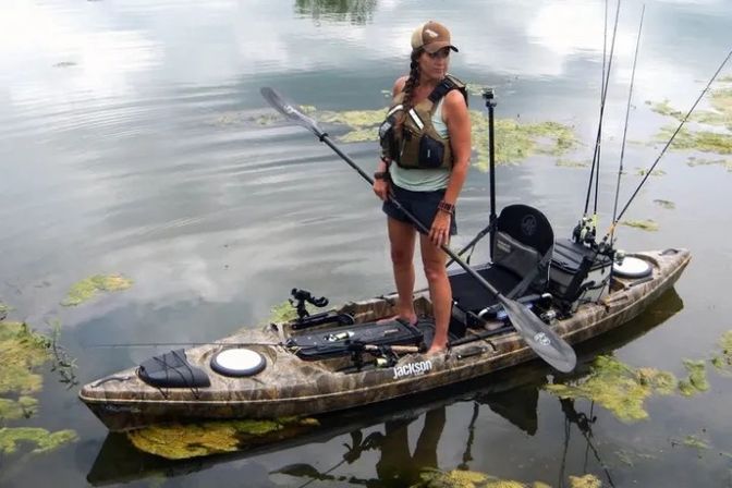 Person standing on a camo fishing kayak in a calm freshwater lake, holding a paddle, wearing a life vest and cap, with mounted fishing rods and floating algae nearby.