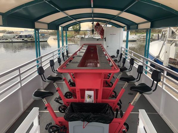 Covered pedal-powered party boat with a long red central bar, swivel saddle seats and cup holders, docked at a marina.