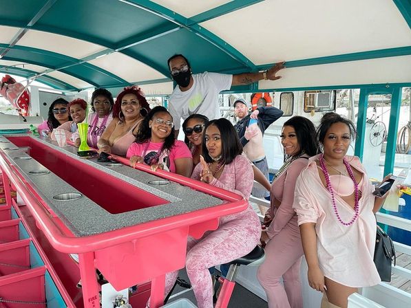 Group of friends smiling and posing at a bright pink bar on a covered party boat, many wearing pink outfits and beaded necklaces, with crew member in a mask and marina scene visible outside.