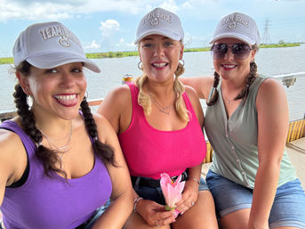 Three friends in white "Team Bride" caps and colorful tank tops smiling on a boat by a marshy waterfront, one holding a pink flower — sunny summer outing.