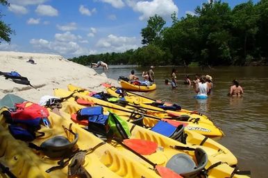 Yellow kayaks lined on a sandy river sandbar with paddles and life jackets, while a group of people wade and swim in a brown river on a sunny summer day under blue sky and green trees.