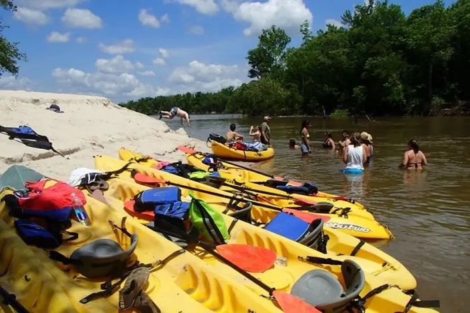 Yellow kayaks lined on a sandy river sandbar with paddles and life jackets, while a group of people wade and swim in a brown river on a sunny summer day under blue sky and green trees.