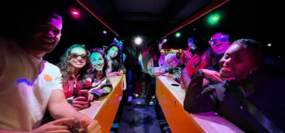 Group of people smiling inside a party bus at night, colorful LED lights, drinks and a long wooden bar creating a lively nightlife vibe