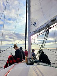 Sunset sail on a sailboat — mast, white sail and rigging framing calm coastal waters as a small, bundled group sits on deck beneath a cloudy sky.