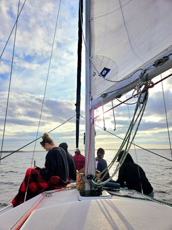 Sunset sail on a sailboat — mast, white sail and rigging framing calm coastal waters as a small, bundled group sits on deck beneath a cloudy sky.