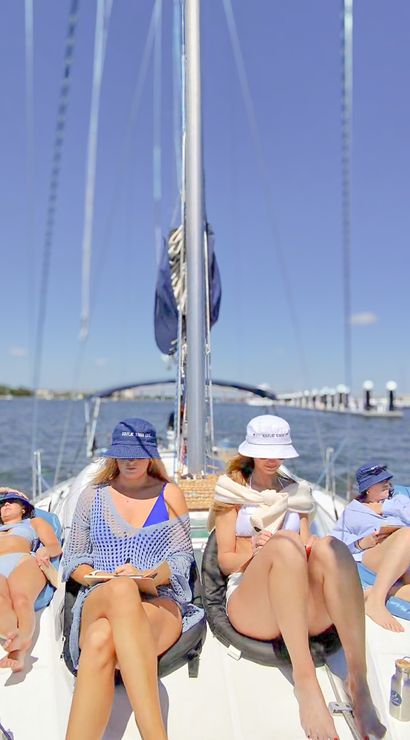 Four people relaxing on the deck of a sailboat on a sunny summer day, lounging in swimsuits and bucket hats with calm harbor docks and blue sky in the background.