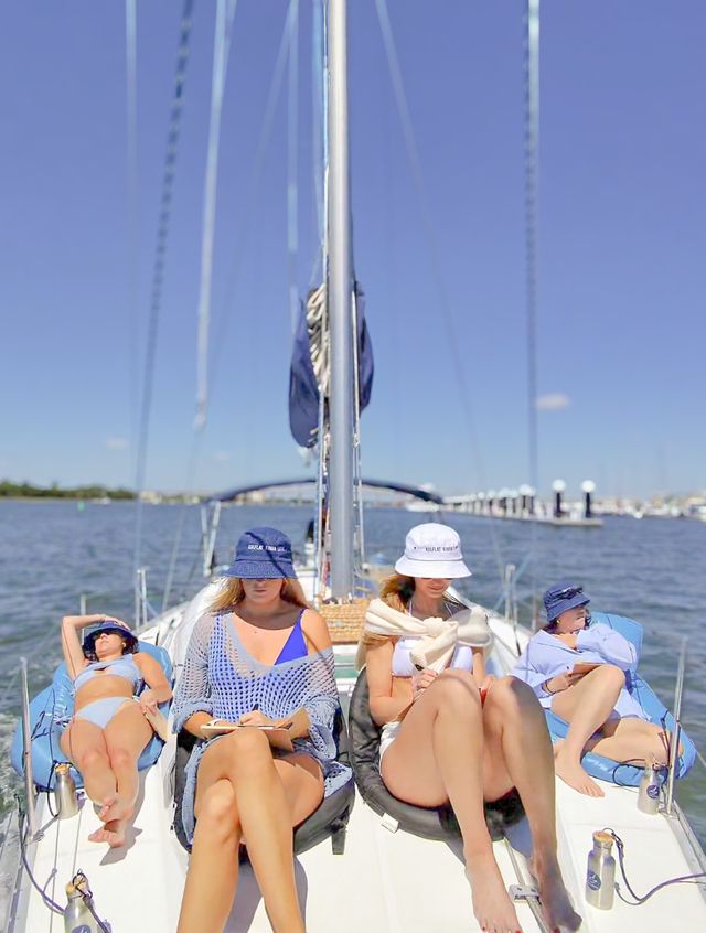 Four people relaxing on the deck of a sailboat on a sunny summer day, lounging in swimsuits and bucket hats with calm harbor docks and blue sky in the background.