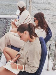 Three people in cozy sweaters and sunglasses writing and relaxing on a sailboat deck with cushioned seats, rigging and calm ocean water in the background