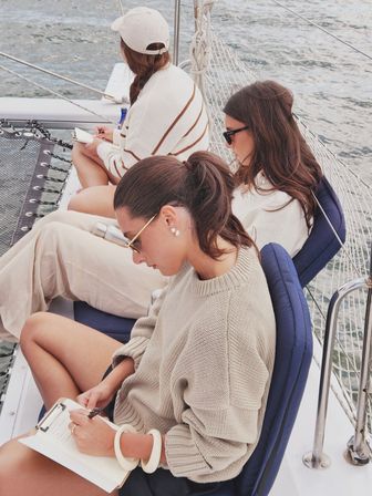 Three people in cozy sweaters and sunglasses writing and relaxing on a sailboat deck with cushioned seats, rigging and calm ocean water in the background