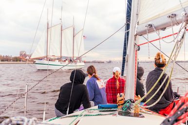 Five people seated on a sailboat bow watching a three-masted schooner with an American flag pass in a coastal harbor under a cloudy sky, with rigging and ropes in the foreground.