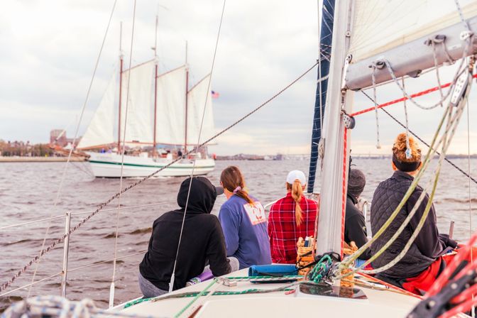 Five people seated on a sailboat bow watching a three-masted schooner with an American flag pass in a coastal harbor under a cloudy sky, with rigging and ropes in the foreground.