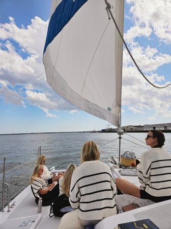 Four women in matching striped sweaters relaxing on a sailboat beneath a large white spinnaker, sailing near a coastal harbor under a blue sky with clouds.