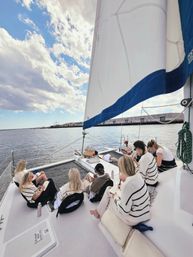 Group of people journaling and relaxing on the deck of a white catamaran sailboat with a large white-and-blue sail, anchored near a coastal harbor under a bright, partly cloudy sky.