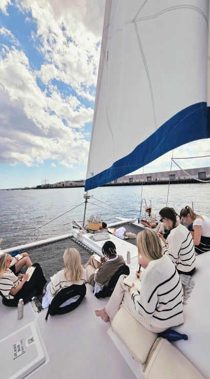 Group of people journaling and relaxing on the deck of a white catamaran sailboat with a large white-and-blue sail, anchored near a coastal harbor under a bright, partly cloudy sky.