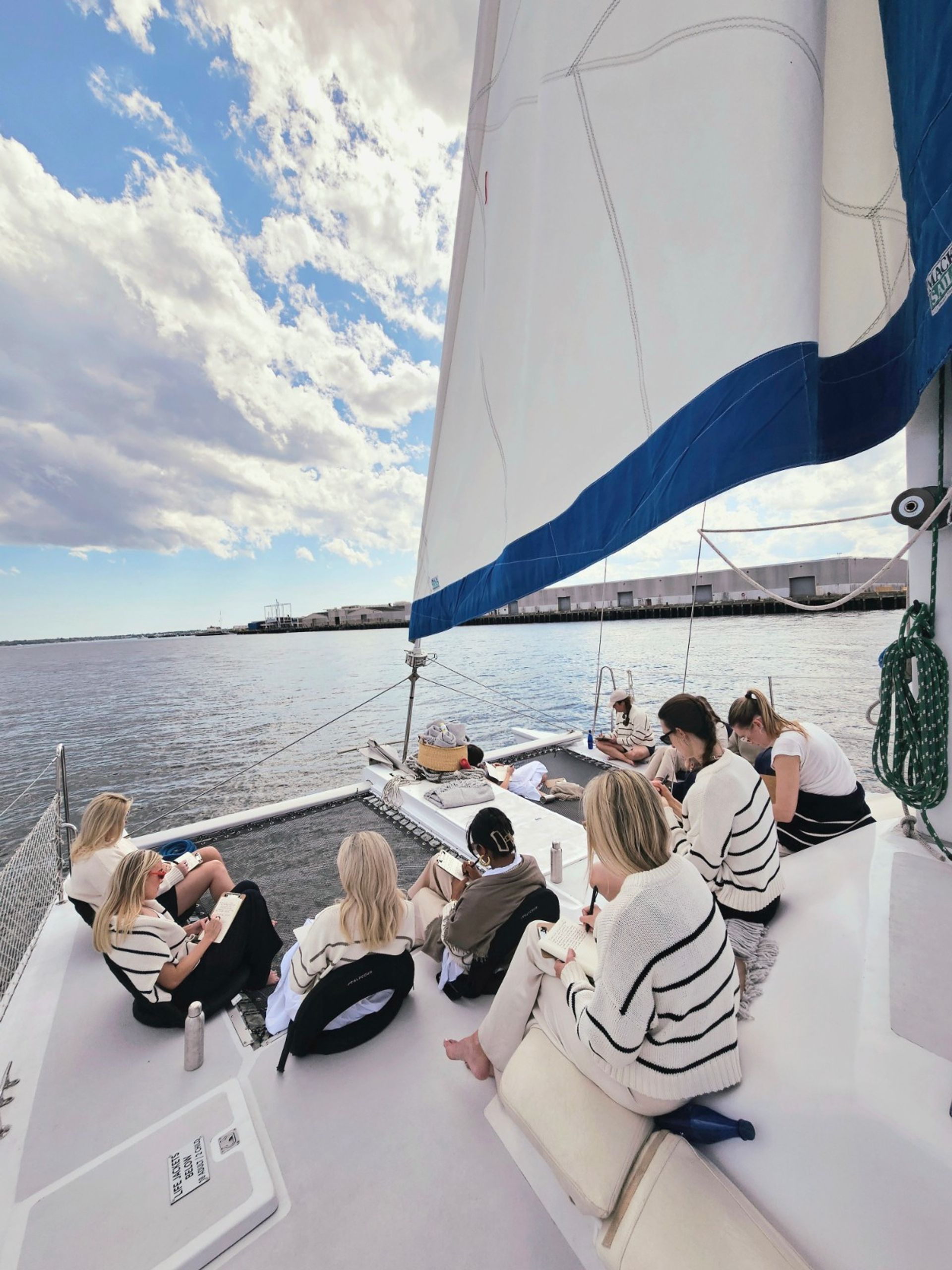 Group of people journaling and relaxing on the deck of a white catamaran sailboat with a large white-and-blue sail, anchored near a coastal harbor under a bright, partly cloudy sky.
