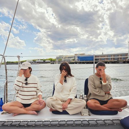 Three people seated cross-legged on a sailboat deck practicing breathing meditation with fingers on their noses, coastal marina and cloudy sky over the harbor in the background.