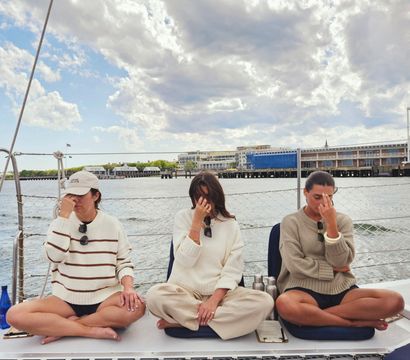 Three people seated cross-legged on a sailboat deck practicing breathing meditation with fingers on their noses, coastal marina and cloudy sky over the harbor in the background.