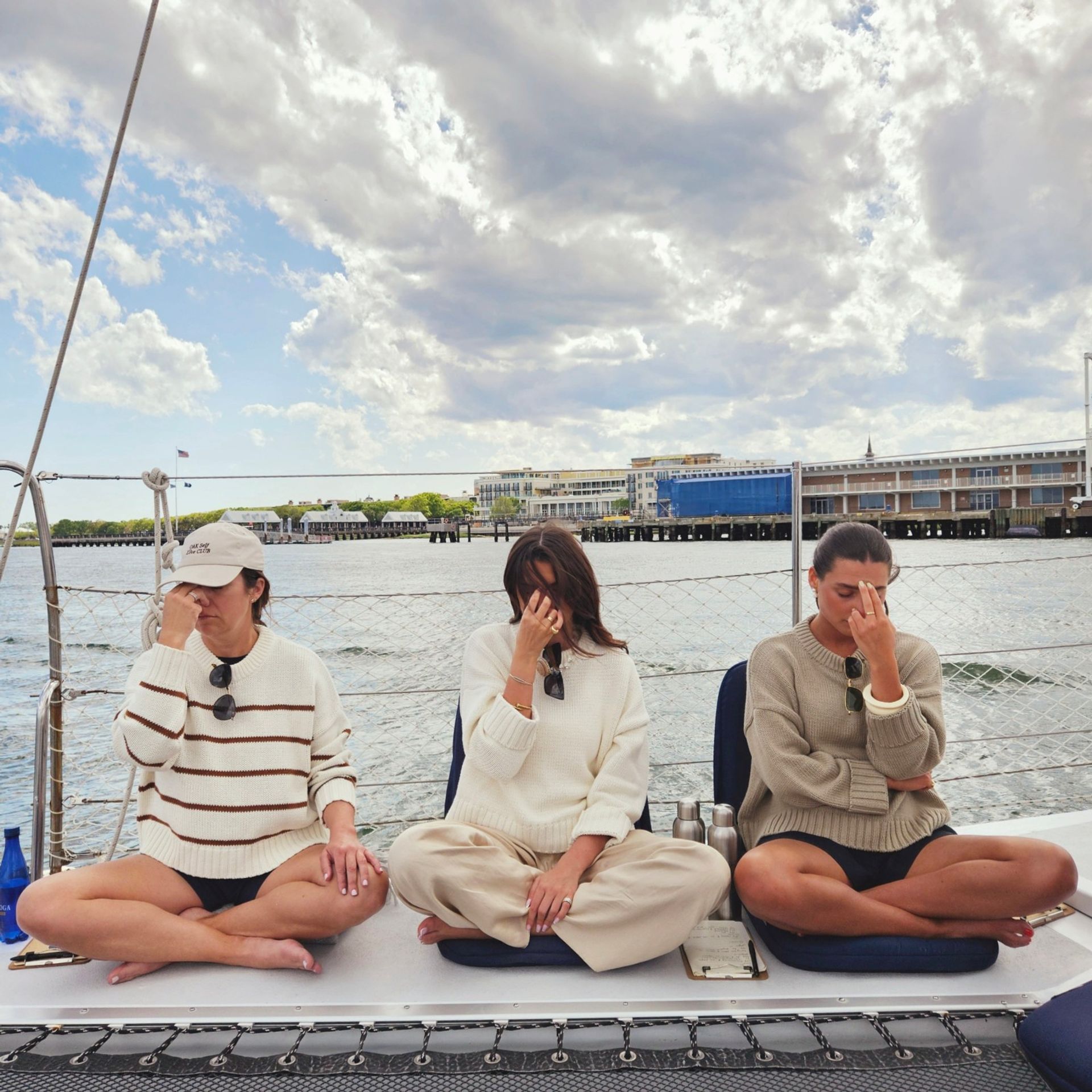 Three people seated cross-legged on a sailboat deck practicing breathing meditation with fingers on their noses, coastal marina and cloudy sky over the harbor in the background.