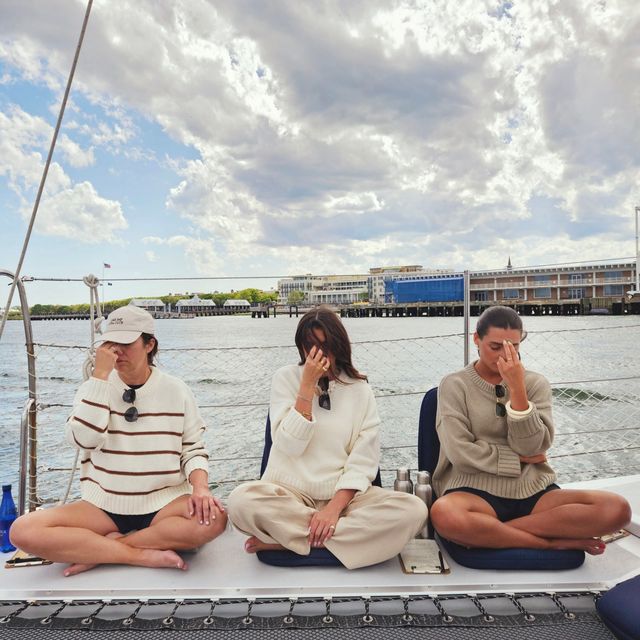Three people seated cross-legged on a sailboat deck practicing breathing meditation with fingers on their noses, coastal marina and cloudy sky over the harbor in the background.