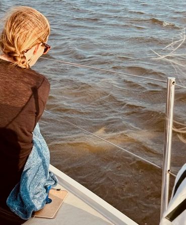 Person with braided hair and sunglasses wrapped in a blue blanket sitting on the bow of a sailboat, leaning on the stainless-steel lifeline and gazing over choppy brown water in warm sunlight.