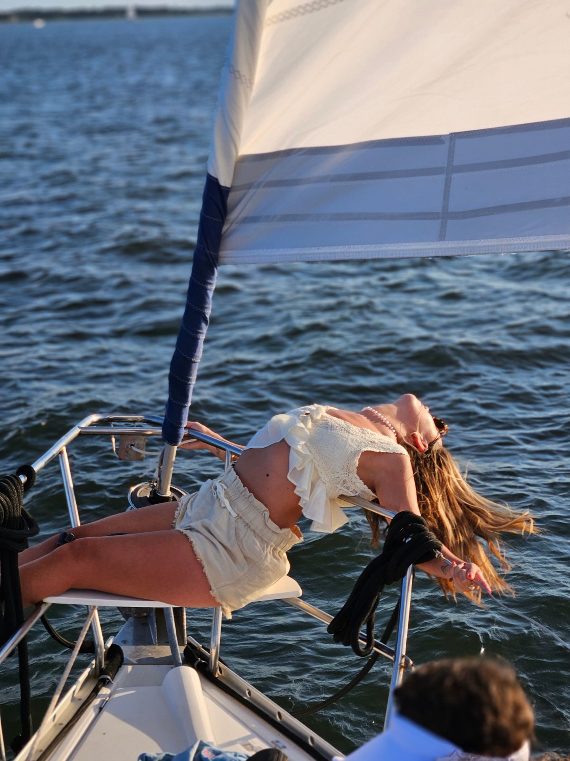 Person reclining on the bow of a sailboat beneath a white sail, sunlit hair and summer outfit with calm blue water and gentle waves in the background — relaxed sailing scene.