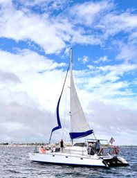 White sailboat with blue-trimmed sails and American flag cruising near a coastal harbor under a bright blue sky with fluffy clouds, people relaxing on deck.