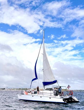 White sailboat with blue-trimmed sails and American flag cruising near a coastal harbor under a bright blue sky with fluffy clouds, people relaxing on deck.