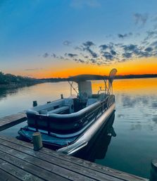 Pontoon boat tied to a wooden dock on a calm lake at sunset, orange sky and scattered cloud reflections.