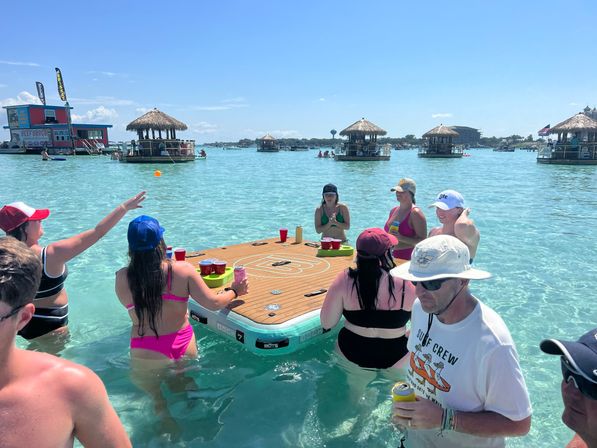 Group of adults in swimsuits standing in clear turquoise shallow water around a floating wooden game table playing a cup-toss game, with thatched tiki huts and boats dotting a sunny tropical lagoon.