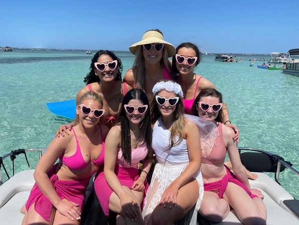 Seven women in pink swimsuits and heart-shaped sunglasses, including a bride in white with a veil, posing on a boat in shallow turquoise coastal water under a sunny blue sky.