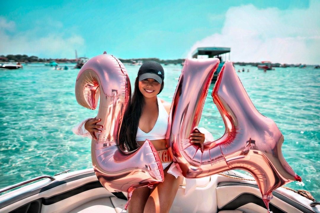 Smiling woman in a white bikini and black cap on a boat holding rose-gold '24' balloons over clear turquoise water at a sunny boat party