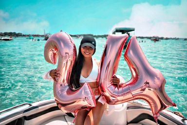 Smiling woman in a white bikini and black cap on a boat holding rose-gold '24' balloons over clear turquoise water at a sunny boat party