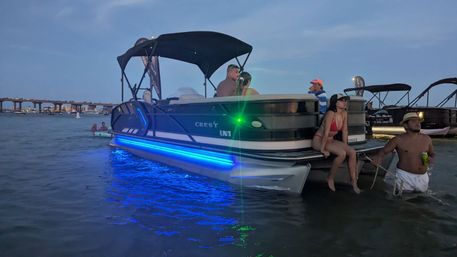 Dusk scene of a pontoon boat with blue LED lights glowing along the hull, anchored in shallow coastal waters near a bridge, people in swimsuits relaxing on the stern and wading beside it with other boats in the background.
