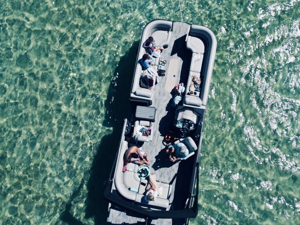 Aerial view of a pontoon boat with people lounging on cushioned seats over clear green water, sunlit ripples and a relaxed summer vibe.