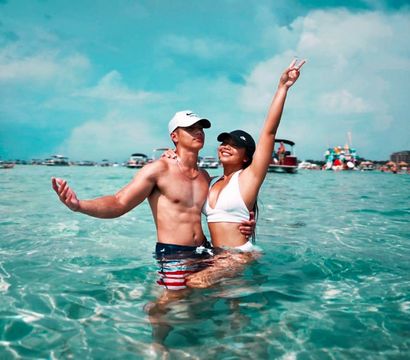 Young couple in clear turquoise ocean near a boat-filled shoreline, woman in a white bikini flashing a peace sign and man in a cap and striped swim trunks enjoying a sunny summer beach day.