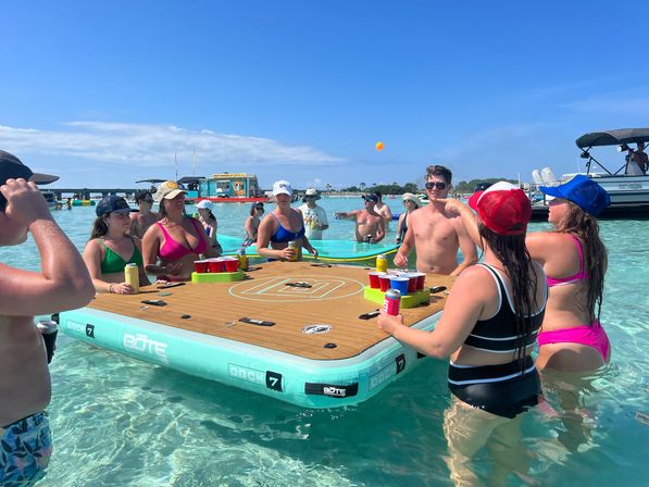 Group of people in swimsuits playing beer-pong on a large inflatable floating table in clear turquoise shallow ocean water near anchored boats under a sunny blue sky